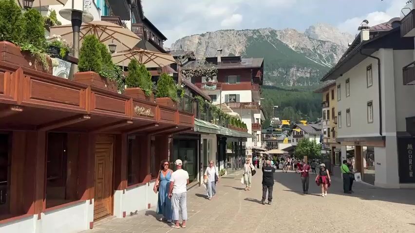 The media file depicts a bustling street scene in Cortina d'Ampezzo, Italy, with the majestic Dolomite mountains serving as a dramatic backdrop. The image captures a sunny day, likely late morning or early afternoon, indicated by the bright lighting and clear skies with some scattered clouds. The weather appears pleasant, encouraging outdoor activity.

The foreground is dominated by the charming architecture of the town. On the left, a multi-story building features a wooden facade with awnings and outdoor seating areas, suggesting cafes or shops. Further down the street, shops with large glass windows are visible. On the right, the buildings are a mix of white and yellow, with balconies and typical alpine architectural elements.

The street itself is paved and appears to be a pedestrian zone, filled with people strolling, shopping, and enjoying the atmosphere. Several individuals are walking in various directions, some in small groups, others alone. They are dressed in casual summer clothing, appropriate for the warm weather. Their body language and the general scene suggest a relaxed and leisurely mood, with a sense of enjoyment of the vacation setting.

Notable details include the lush greenery planted in pots and along the buildings, adding to the picturesque ambiance. The distant mountains are rugged and imposing, their rocky faces catching the sunlight. The overall impression is one of a vibrant, attractive tourist destination.

Visible text includes "MOORE" on a sign on the right side of the street, likely indicating a business name. There might be other smaller signs on shopfronts, but they are not clearly legible. The scene conveys a strong sense of place, a European mountain resort town alive with activity and natural beauty.