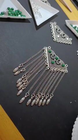 A video shows a close-up, top-down view of jewelry-making supplies on a black surface. Several ornate silver pendant bases are laid out, some with decorative swirls and small holes for attachment. One larger pendant, embellished with small green beads, hangs down with multiple chains, each ending in a silver tassel-like charm. Small white triangular dishes hold tiny green beads and clear beads. In the background, other jewelry components and crafting tools are visible. The lighting is bright, illuminating the details of the jewelry pieces.