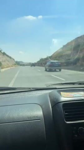 The video shows the point of view of someone driving a car on a highway. The driver, a man wearing a watch, is visible in the frame. Other cars are also visible on the road. The dashboard of the car is prominently displayed, with an air freshener hanging from the rearview mirror. The surroundings consist of a clear blue sky and hillsides with some vegetation along the highway. The video appears to be taken during daylight hours.