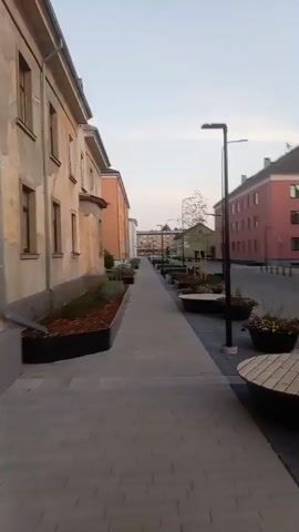 The video shows a person walking down a paved pathway lined with raised garden beds on either side. The garden beds contain mulch and some sparse vegetation. On the left, there is a light-colored building with visible signs of wear and tear. On the right, there are modern buildings painted in vibrant colors, including orange and pink. The pathway leads towards a distant intersection with more buildings and streetlights. The sky is overcast and the lighting suggests it is either early morning or late evening. A person is briefly visible on the left, standing with a bicycle. The overall scene depicts an urban or semi-urban environment that appears to be undergoing redevelopment or has recently been improved with new landscaping and seating areas.