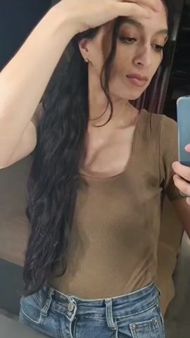 A woman is in a dressing room, taking a selfie video. She is wearing a brown, short-sleeved, square-necked t-shirt and blue jeans. Her long, dark hair is pulled to one side. She is holding a blue smartphone in her right hand and occasionally touches her hair with her left hand. The background shows parts of the dressing room, including a mirror, and a curtain.