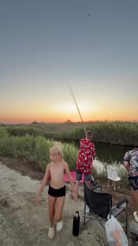 The video shows a sunset scene at a body of water. In the foreground, a young girl with blonde hair and wearing dark shorts stands to the left. To the right, a boy wearing a red floral shirt and dark shorts is fishing. He holds a fishing rod and casts it into the water. Behind him, another person is partially visible. Several chairs and a water bottle are on the ground. The sky is a gradient of orange and blue, with tall grass lining the water's edge.