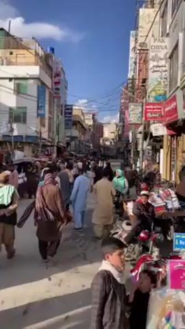 A busy street in an urban area is shown in the video. Many people, including men, women, and children, are walking along the street. Some of the men are wearing traditional clothing, while others are wearing more modern attire. There are also several motorcycles parked on the side of the street. A small table of goods, likely for sale, stands on the right of the street. The buildings on either side of the street are a mix of old and new, and there are signs and banners hanging from some of them. The sky is blue and there are some clouds in the distance. The overall impression is of a bustling, lively place.