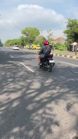 The video shows a daytime scene of a two-lane asphalt road with trees and buildings lining the right side. In the foreground, shadows of leaves are cast on the road. Two people are riding a black motorcycle in the right lane, heading away from the camera. The person in front is wearing a red helmet and a grey shirt, while the person in the back is wearing a black shirt. Cars and trucks are visible further down the road, also traveling in the same direction. Power lines and poles are present along the roadside. The sky is blue with scattered clouds.