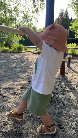 A young child wearing a brown hat with protruding ears, a white t-shirt, green shorts, and brown sandals hangs from a horizontal bar outdoors on a playground. The child is facing away from the camera, gripping the bar with both hands. The child’s legs dangle below. The bar is secured between two blue metal poles. The ground underneath the bar is covered in gray gravel. Green trees and shrubbery are visible in the background behind the playground equipment.