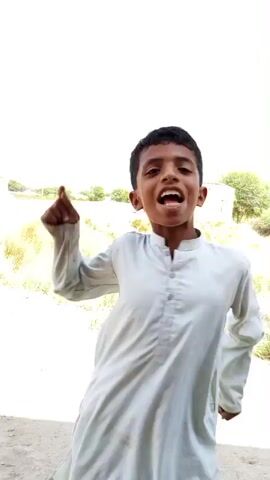 The video shows a young boy in a light-colored kurta and shalwar standing in an outdoor environment. The boy is laughing and appears to be looking at the camera. In the background, there is a building with columns, some dry vegetation, and a clear sky. The boy moves his arms and dances playfully. The camera is close to the boy's face at times, capturing his expression. The scene suggests a rural or semi-rural setting.