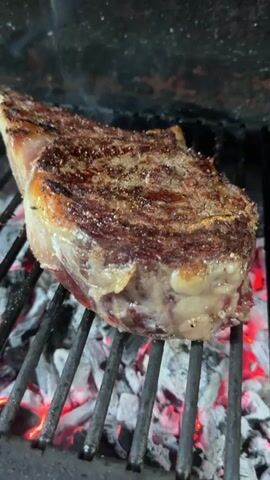 The video shows a thick cut of steak, likely a ribeye, being grilled over hot coals. The steak has a crust forming on the outside, with grill marks visible. The coals are glowing red, indicating high heat. The steak is positioned on a metal grill grate. The background is dark, suggesting an outdoor grill or a dark kitchen setting. The camera is focused on the steak, with a slight zoom in and out throughout the short clip.