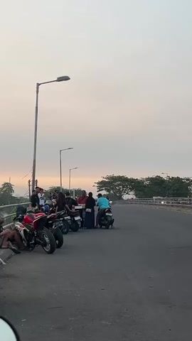 A group of people and several motorcycles are gathered on the side of a road. A man in a blue shirt is riding a scooter. There are trees in the background and the sky is overcast.