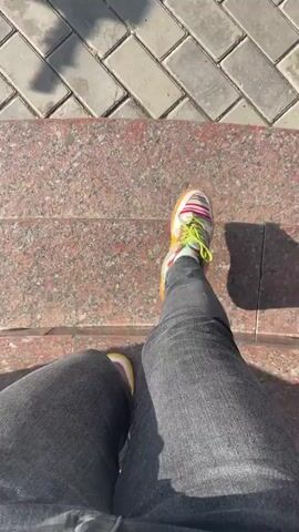 This video shows a person walking on a paved sidewalk. The camera angle is from the person's perspective, looking down at their feet and the ground ahead. The person is wearing dark jeans and colorful sneakers. The sidewalk is made of rectangular gray pavers. In the background, there are cars parked on the street, trees, and a tall apartment building. The sky is blue with some white clouds, suggesting it is daytime and the weather is fair. The scene appears to be in a residential or urban area. The person is walking forward at a steady pace. There are shadows cast on the ground from an unseen object, possibly a tree or a building. The video does not contain any visible text. The overall mood is casual and ordinary, depicting a simple walk.