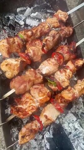 Four skewers of meat and vegetables are grilling over charcoal. The meat appears to be chicken or pork and is cubed. Red bell peppers and green peppers are interspersed between the meat on the skewers. The meat is browning and some pieces are showing char marks. Smoke is rising from the grill.