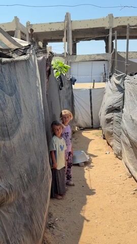 Two young girls stand in a narrow dirt path between makeshift shelters constructed from dark tarpaulin. The girl on the left wears a light-colored t-shirt with a blue butterfly graphic and a long, patterned skirt. Her dark hair is pulled back. The girl on the right wears a light purple t-shirt with a floral pattern and matching patterned trousers. Her blonde hair is loose. Both girls have fair skin and appear to be looking towards the camera. The shelter walls are made of draped fabric and ropes. Behind the shelters, a partially constructed concrete building with exposed rebar and visible damage stands under a clear blue sky. A silver basin rests on the ground near the girls. The ground is dry, sandy soil.