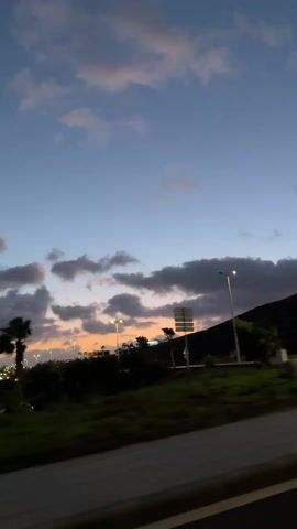 The video appears to be shot from a moving vehicle, likely a car, at dusk or twilight. The sky is a gradient of blue and orange with scattered clouds. Silhouetted palm trees line the roadside. The camera passes by buildings, one of which has a lit window emitting a warm yellow light. Streetlights are visible in the distance, adding to the artificial illumination. The road and a partial view of the vehicle's interior can be seen in the foreground. The camera movement is slightly shaky, typical of a hand-held recording.