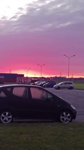 The video shows a parking lot with a vibrant pink and purple sunset. Several cars are parked in the lot, including a black hatchback in the foreground. Shipping containers and streetlights are visible in the background. The sky is filled with clouds, and the sun appears to be setting on the horizon.