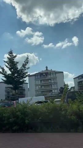 The video pans across a street view of a residential area with apartment buildings under a partially cloudy sky. Cars are parked along the street, and some people are visible walking near the parked vehicles. There are trees and bushes in the foreground, and a red car is prominent in the shot. A bus stop sign is visible.