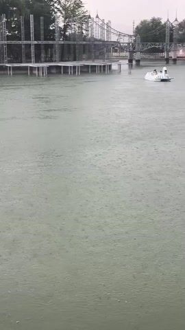 The video shows a body of water, likely a lake or river, during a rain shower. The water's surface is covered with ripples and splashes from the falling rain. In the background, a man-made structure, possibly a pier or boardwalk, is visible extending into the water. Beyond that, there are trees and a bridge or overpass. A small boat with a few people on board is visible on the water in the middle ground, navigating through the rain. The overall atmosphere is one of a quiet, overcast day with steady rainfall.