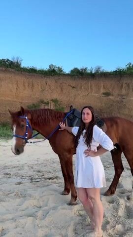 The video shows a woman standing next to a brown horse on a beach. The woman is wearing a short, white dress. She has long, dark hair. The horse is saddled and has a blue bridle. There is a sandy cliff in the background. The sky is a clear blue. A man is visible in the background, near a white horse. The woman poses, then pets the horse. The horse shakes its head.