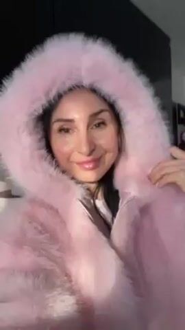 A woman with brown hair is standing indoors wearing a pink faux-fur coat with a large hood. She smiles and looks directly at the camera. The background includes a black wall and a blurred interior of a home. Sunlight shines on her face.