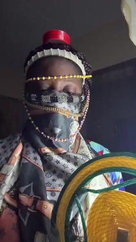 A person stands indoors, facing the camera. They are wearing multiple headbands, beaded jewelry around their eyes, a patterned face mask, and a scarf. They also have a red object balanced on their head. They are wearing a colorful patterned jacket or shirt. They move a yellow object in front of the camera. The background is dimly lit, with hints of a window and curtains.