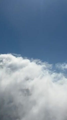 This video shows a view from an aircraft looking down at a layer of clouds. The sky above is a clear blue. The clouds are white and fluffy, with some areas appearing darker and more textured, suggesting depth and possibly some thicker cloud formations. The camera appears to be moving forward, as the perspective of the clouds changes slightly. There are no people or objects visible other than the clouds and the sky. The overall impression is one of soaring above the clouds in a bright, clear day.