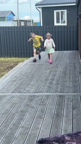 Two young children are on a wooden deck. A boy wearing a yellow shirt with a graphic and patterned shorts is in the foreground on the left, appearing to be running or dancing. A younger girl with blonde hair, wearing a white long-sleeved shirt with text and a patterned skirt, is on the right. She is looking at the boy and appears to be talking. The background shows a dark fence and a building. The overall environment suggests an outdoor setting, possibly a backyard.