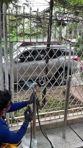 A person is welding a metal pole in front of a decorative metal fence. The person is wearing a blue shirt and black gloves. They are holding a welding torch and sparks are flying. A silver SUV is parked behind the fence. The environment appears to be outdoors.