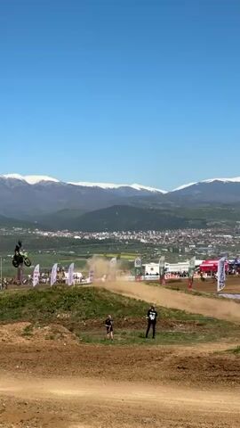 The video shows a motocross race. A rider on a dirt bike is seen performing a jump over a small hill on a dirt track. The landscape in the background features a town and mountains with snow-capped peaks under a clear blue sky. Several flags are seen alongside the track. Two people, a child and an adult, are observing the race from the side of the track.