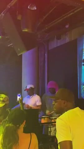 The video shows a night club setting. In the foreground, there are people with various hairstyles and clothing. At the center, a musician is playing the congas, wearing a pink wig. To his left, another man is standing at a DJ setup, holding his phone up to record. Behind the crowd, a screen displays text including "Jyde Ajala" and "lo see." The lighting in the club is dynamic, shifting between purple, green, and yellow, adding to the vibrant atmosphere. Some people are standing, while others are seated on a couch. The overall scene suggests an energetic performance in a crowded club.