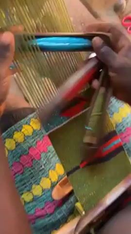 A person is weaving a piece of fabric with an intricate design using a hand loom. The fabric features a pattern of colorful geometric shapes, including diamonds and trapezoids in shades of green, blue, red, black, and yellow. The weaver uses a variety of tools, including shuttles holding different colors of yarn. The person's hands are visible as they manipulate the loom, inserting and securing the yarn. In the background, there are additional items, including scissors and a pair of sandals, placed on a wooden surface. The scene is shot from a high angle, giving a clear view of the weaving process and the materials involved.