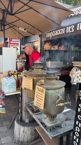A street vendor is serving tea from a samovar to customers. The vendor is an older man wearing a hat and a red vest. He is standing behind a table laden with jars of honey, bottles of water, and other goods. Two customers are visible, a woman in a green and white striped shirt and another woman in a black shirt. The samovar is made of brass and is steaming. There are signs on the samovar with Russian writing. The scene appears to be at an outdoor market.