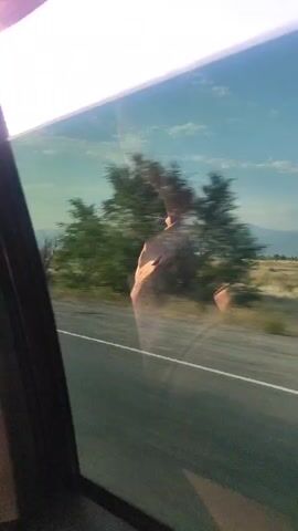 The video captures a view from inside a vehicle looking out the passenger side window. The window reflects a person, likely the videographer, with their right arm extended upwards and their hand visible. Outside the window, a rural landscape is visible, with a road, sparse vegetation, trees, and distant mountains under a bright sky. The sunlight creates a glare on the window, partially obscuring the view. The camera is moving, indicated by the motion blur of the passing scenery.
