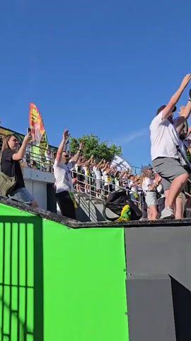A group of people are on a raised platform, possibly a stage or float, under a clear blue sky. They are waving flags and arms in celebration. The person on the far left is a woman wearing a black t-shirt and shorts, holding a large colorful flag. Next to her, a man in a white t-shirt and black pants is also raising his arms. In the background, many people in white t-shirts are gathered, some holding white flags. On the right side of the platform, a man in a white t-shirt and grey shorts is visible, along with a child in a white t-shirt and shorts. A black bag with a yellow and green object is placed on the platform. The platform has a vibrant green side with black accents.
