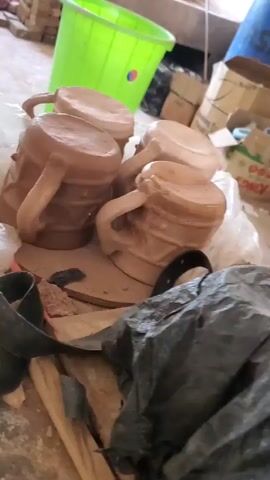 The video shows a group of unglazed clay mugs on a table in a cluttered workshop environment. The mugs are light brown, cylindrical in shape, and have handles. They are partially covered with a crumpled plastic bag. A long, curved, dark-colored tool lies nearby. The background includes a green plastic bucket, cardboard boxes, and various other unidentified items.  The lighting is somewhat dim, and the camera moves slightly, providing different angles of the mugs.