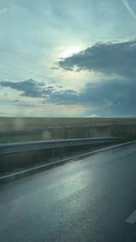 The video captures a view from a vehicle moving along a wet asphalt road. The sky is partly cloudy with the sun visible through some of the clouds. The landscape features a field with some vegetation, including what appear to be red flowers, and trees. In the distance, there are power lines and a faint haze. The road surface reflects the sky and the sunlight, indicating it is wet, possibly from recent rain. There are no people or distinct interactions visible. The overall atmosphere is serene and suggests travel through a rural or open area.
