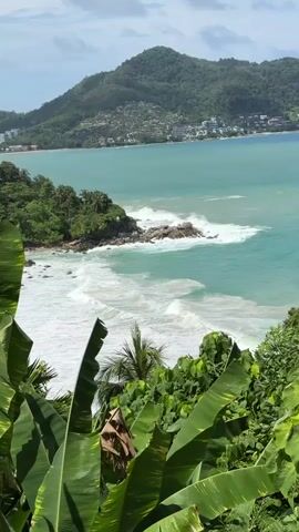 This video captures a scenic view of a tropical coastline. In the foreground, lush green foliage, including large banana leaves, partially obstructs the view. Beyond the foliage, the turquoise ocean stretches out, with white waves crashing against rocky outcrops and a small, tree-covered island. In the distance, a bay is visible, lined with buildings and nestled at the base of verdant, tree-covered hills. The sky above is a mix of blue and scattered clouds. The camera appears to be positioned at an elevated vantage point, looking down towards the sea and the distant coastline. There are no people or animals visible in the video. The overall scene is one of natural beauty and tranquility.