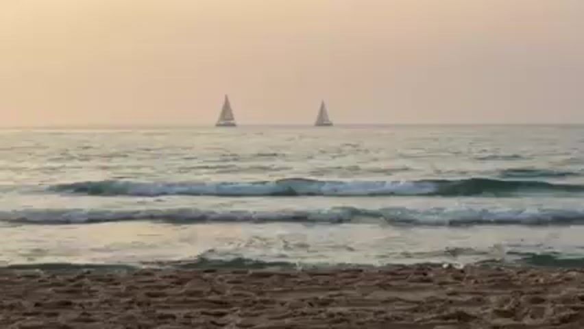 The video shows a serene ocean view from a sandy beach. Two sailboats are visible on the horizon under a pale, slightly hazy sky. Gentle waves roll onto the shore. The beach is composed of brown sand.