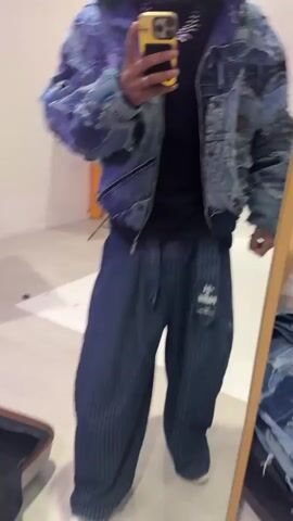 The video shows a young man posing in front of a mirror, showing off his outfit. He's wearing a denim patchwork jacket with a hood, a black t-shirt, loose pinstripe pants, and white sneakers. He is taking a selfie with his phone. The camera pans around to show a room that appears to be a photography studio. There are several other people in the background, along with lighting equipment, backdrops, and clothes racks. He takes off his denim jacket to reveal a white t-shirt and a second denim jacket.