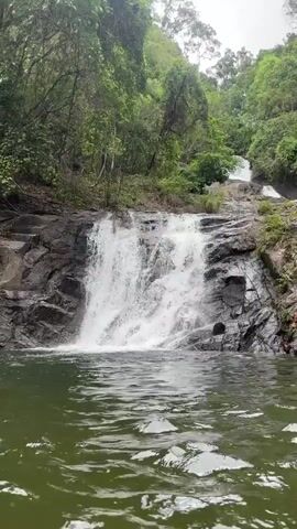 The video shows a waterfall cascading down a rocky slope into a pool of water. The waterfall is the main focus, with white water flowing over dark gray rocks. Lush green vegetation surrounds the waterfall, indicating a natural, possibly forested environment. The camera angle provides a clear view of the waterfall and its immediate surroundings. The overall tone is peaceful and serene.