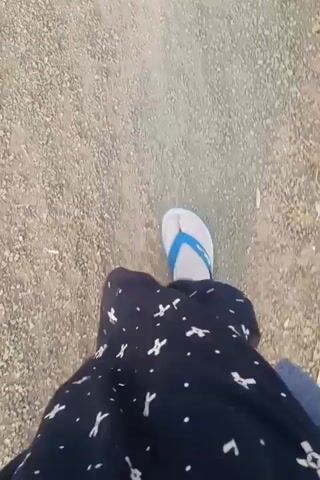 The video shows a first-person perspective of someone walking on a gravel path. The person is wearing dark pants with a white pattern and white flip-flops. The camera angle is looking down at the person's legs and feet as they walk forward. The environment appears to be outdoors with dry, loose gravel on the ground.