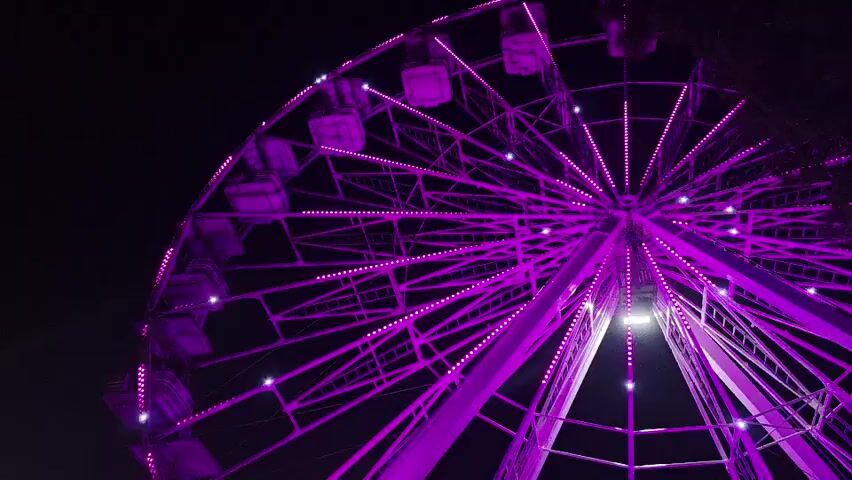 This is a low-angle, close-up shot of a large Ferris wheel at night. The Ferris wheel is illuminated with purple lights, and the lights on the spokes and around the passenger cabins are visible. The sky behind the Ferris wheel is dark. The Ferris wheel appears to be stationary.