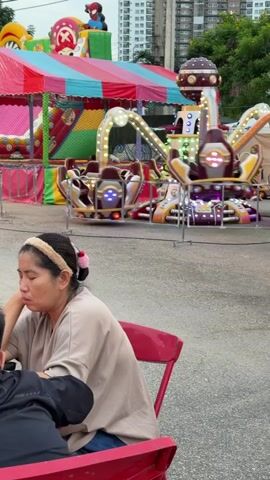 A woman with her eyes closed and head tilted down sits at a red table in the foreground. She is wearing a beige shirt and a light brown headband. In the background, an amusement park ride with spinning seats and lights is in motion, and a colorful inflatable bounce house is visible. The ground is paved, and there are buildings in the distance.