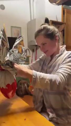 A young woman wearing a plaid pajama shirt is sitting at a wooden table. She is unwrapping a large, Kinder-branded chocolate Easter egg wrapped in foil and decorative paper. She pulls at the foil and paper, slowly opening the egg. The kitchen in the background contains a gas water heater, wooden cabinets, and various household items.