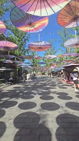 A bustling street market is depicted from a low-angle perspective, looking down a pedestrian walkway. The path is lined with shops on both sides, their awnings and stalls visible. Overhead, a canopy of colorful, decorated umbrellas is suspended, casting circular shadows on the paved ground. Several people are walking along the street. On the left, a few individuals are seated at outdoor tables. The scene is illuminated by bright sunlight, suggesting it is daytime. Trees with green foliage flank the sides of the walkway.