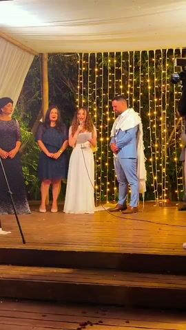 The video captures a wedding ceremony taking place outdoors under a white canopy. A wooden platform serves as the stage, adorned with string lights behind the participants. On the platform, four individuals are present. On the far left, a woman in a dark, sequined dress and a head covering stands facing the center. Next to her, a woman in a dark blue, knee-length dress also faces the center. In the middle, a woman in a white wedding dress and veil is speaking into a microphone, holding a piece of paper. To her right, a man in a light blue suit, wearing a tallit (Jewish prayer shawl) over his shoulders, stands facing the woman speaking. Another person, partially visible on the far right, appears to be operating a camera. The atmosphere suggests a formal occasion, likely a religious ceremony.