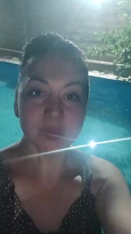 The video shows a woman in a polka dot swimsuit standing next to a swimming pool at night. She is looking at the camera and smiling. The lighting is dim, with a bright light reflecting off the water. The background is blurred, but appears to be an outdoor setting with some greenery.
