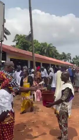 The video appears to depict an outdoor gathering or event, possibly a religious service or a community celebration, taking place in what looks like a field or open area. There are many people in attendance, some wearing traditional or formal attire. Individuals are shown collecting money or donations using a basket. A covered stage or platform is set up, potentially for speakers or performers. The environment includes trees, buildings, and an ambulance on standby, suggesting a well-organized event with safety measures in place. Some people are walking around, carrying water bottles. Overall, the scene conveys a sense of community involvement and organized activity.