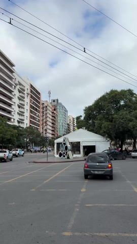 The video shows a street scene in an urban environment, likely in Argentina, judging by the flags being carried by a group of people marching down the street. Tall buildings line both sides of the road. Traffic signals are visible at intervals, all showing red lights. A white tent is erected in the middle of a parking area marked with yellow lines. A sign or structure is in front of the tent, its details difficult to discern. Two white pickup trucks with markings "TRÁNSITO" (Transit) and police lights are escorting the marching group. The sky is overcast.