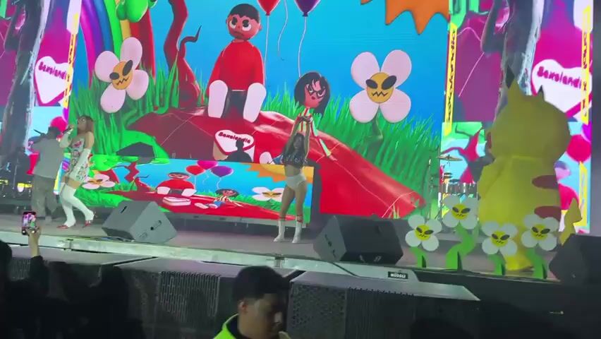 A stage performance with a large screen displaying colorful animated graphics. The screen features oversized cartoon characters, flowers, and a rainbow. On stage, several performers are visible. One performer, dressed in a white outfit with colorful patterns and white boots, is singing into a microphone and dancing. Another performer, wearing a light-colored outfit, is also dancing. Further back, two figures are seen at DJ turntables. To the right of the stage, a large inflatable Pikachu character stands. In the foreground, the audience is partially visible, with one person holding up a phone to record the performance. The stage is lit with spotlights and smoke effects are present. The overall atmosphere appears to be a lively concert or festival.