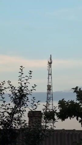 The media file shows a telecommunications tower silhouetted against a pale blue sky with scattered clouds. The sky suggests it's either early morning or late evening. The tower is a lattice structure with several antennas and what appear to be equipment boxes attached. In the foreground, the dark silhouettes of tree branches with leaves and a brick chimney are visible, framing the tower. The lower portion of the image shows the top of a grey roof or wall. The scene is outdoors, likely in a residential or semi-rural area given the presence of trees and a chimney. There are no people visible, and the overall mood is tranquil and somewhat stark due to the silhouettes. The time of day is suggested by the soft, diffused light and the colors in the sky. The location context of Izmail, Ukraine, places the scene in Eastern Europe. There is no visible text in the media file.