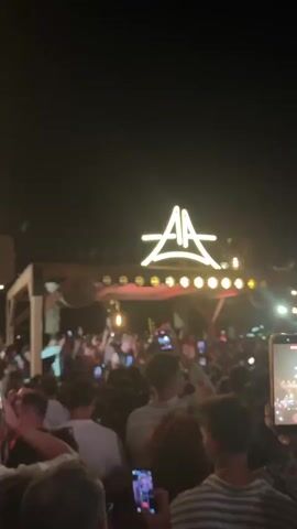 The video shows a crowded outdoor event at night. A large, illuminated sign with the letters "AA" is visible above a wooden structure, possibly a stage or bar, adorned with string lights. A dense crowd of people is gathered below, many holding up their phones. Some individuals are dancing or raising their hands in the air, indicating a lively atmosphere, likely a concert or party. The lighting is dim, with the primary illumination coming from the sign and the string lights, creating a festive ambiance. The overall impression is one of a vibrant, nocturnal gathering.