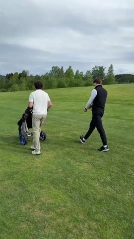 Two men are on a golf course. One man, wearing a white shirt and tan pants, is pulling a golf cart. The other man, wearing a white shirt, black vest, and black pants, is walking beside him. The golf course is green and grassy, with trees in the background. The sky is cloudy.